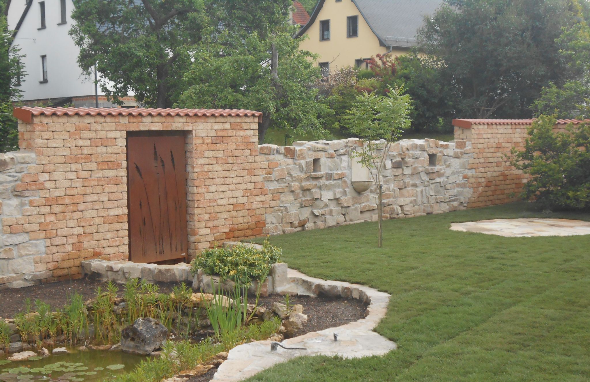 Natursteinmauern mit Ziegelabdeckung und Cortenstahl-Tür, davor Teich und Rasenfläche